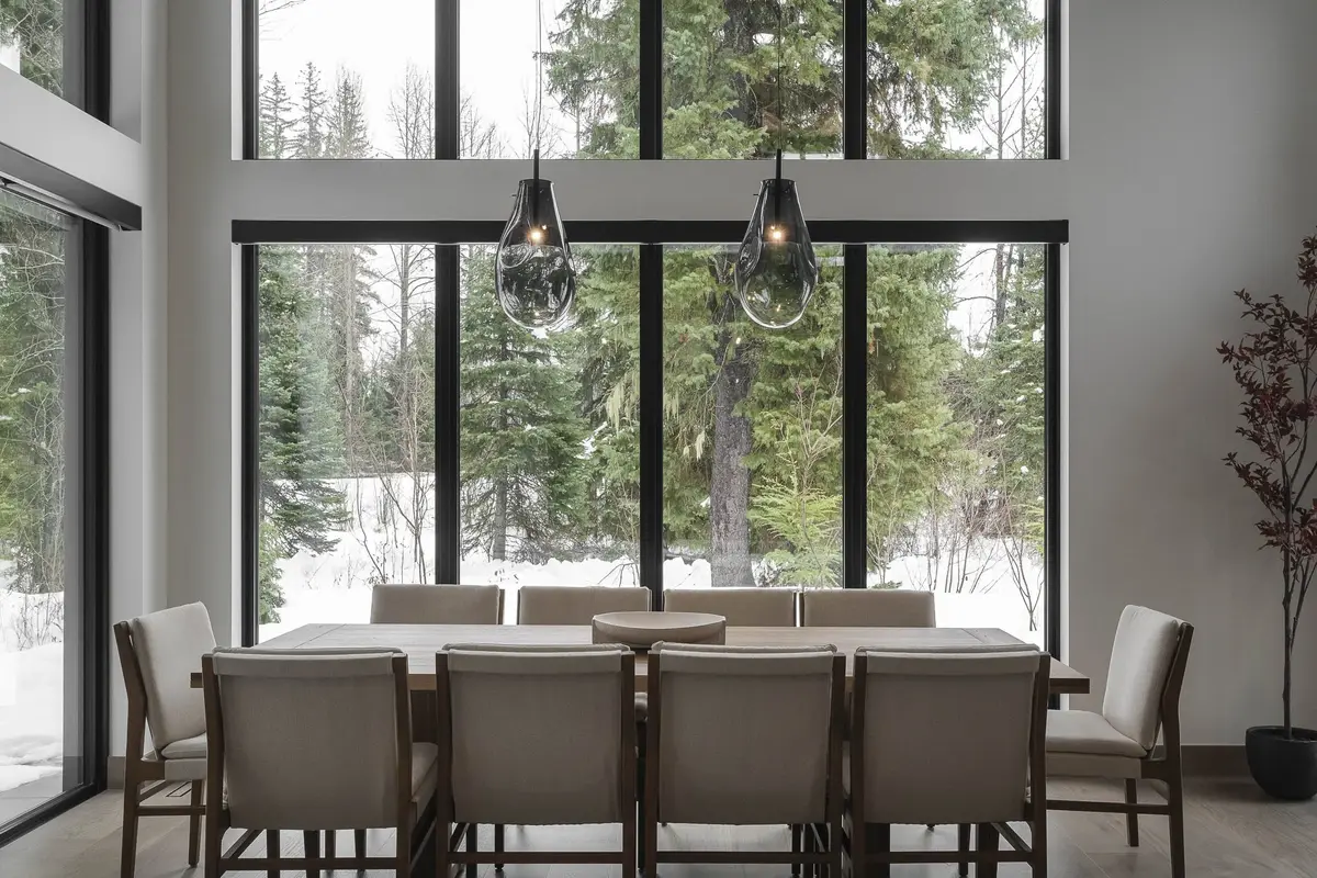 Dining table and chairs under two glass pendants, full height glazing to snow and conifers