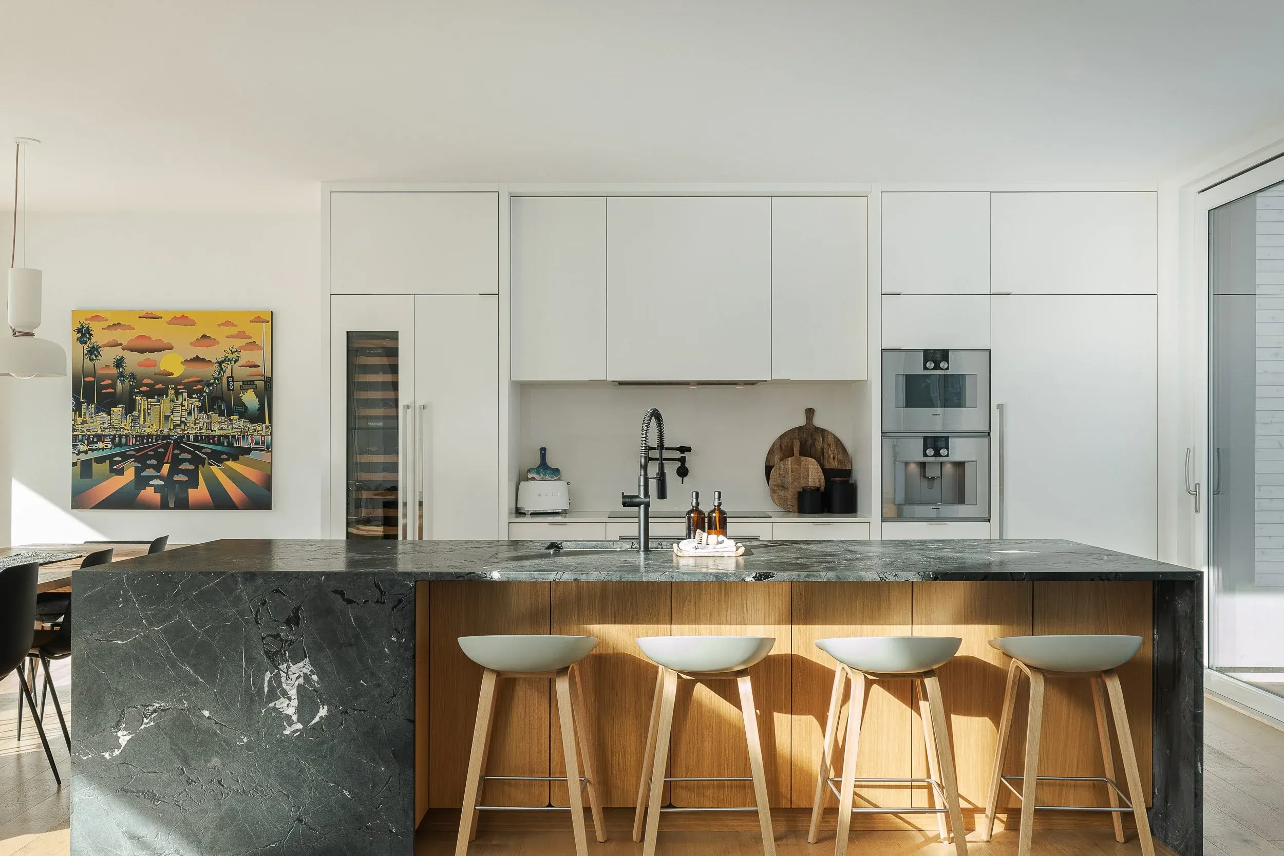 Kitchen island in dark veined stone above wood fronts, four stools, pale cabinetry behind