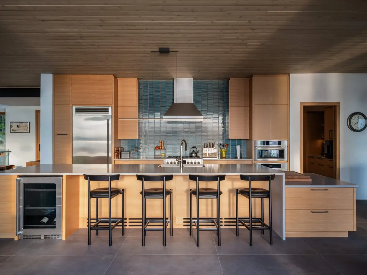 Kitchen island with four stools under a wood ceiling, ovens set into the cabinetry