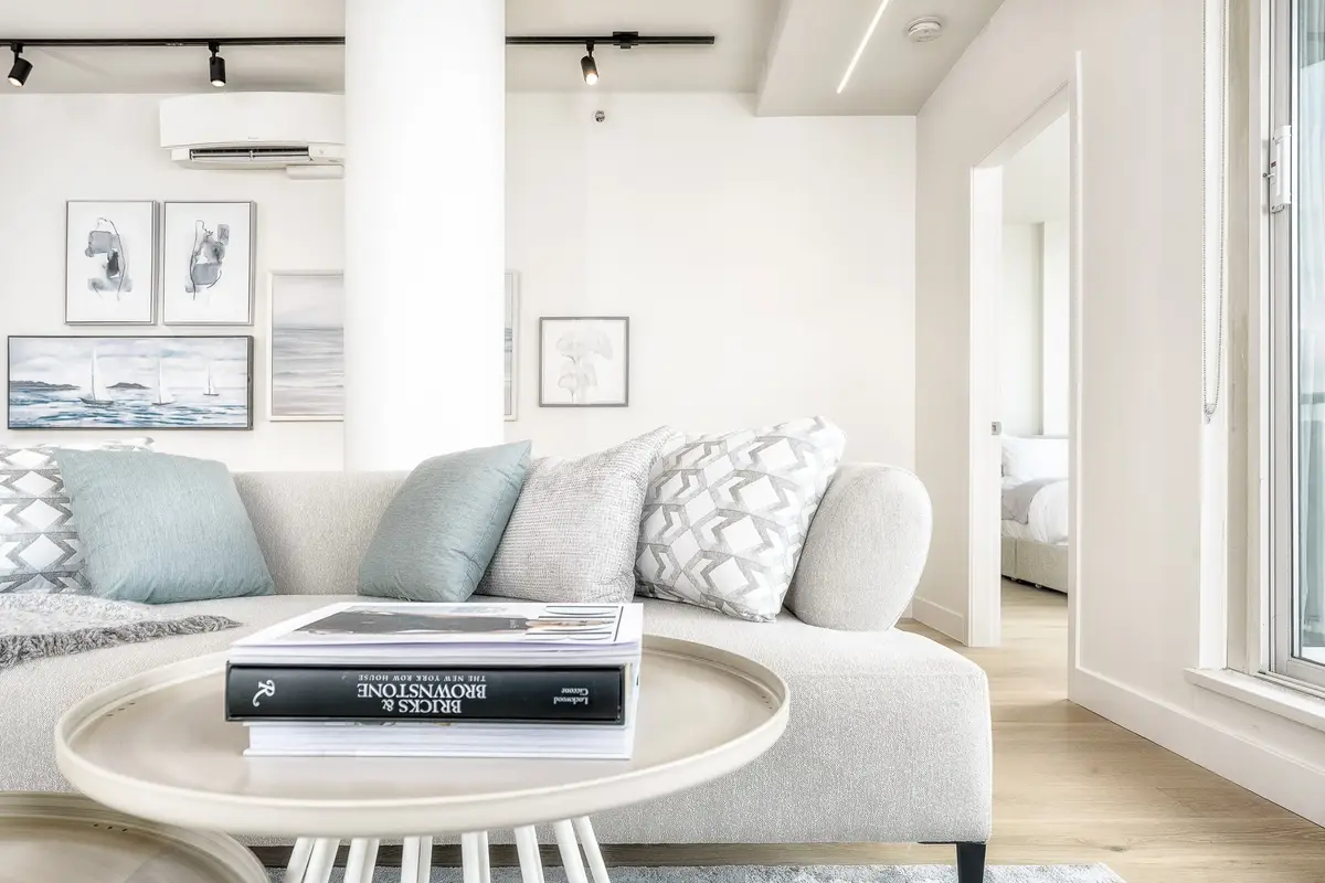 Pale sectional and a round table stacked with books, framed prints above and a bedroom beyond