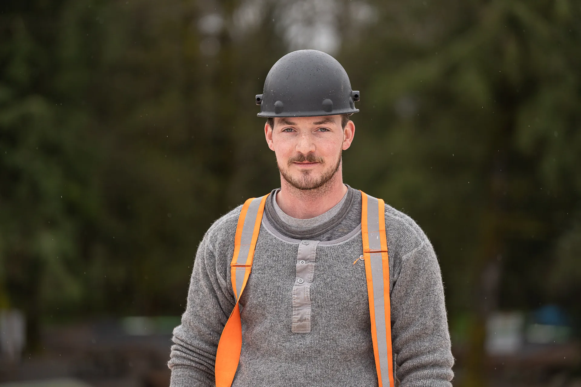 A builder in a hard hat and orange suspenders, trees behind him