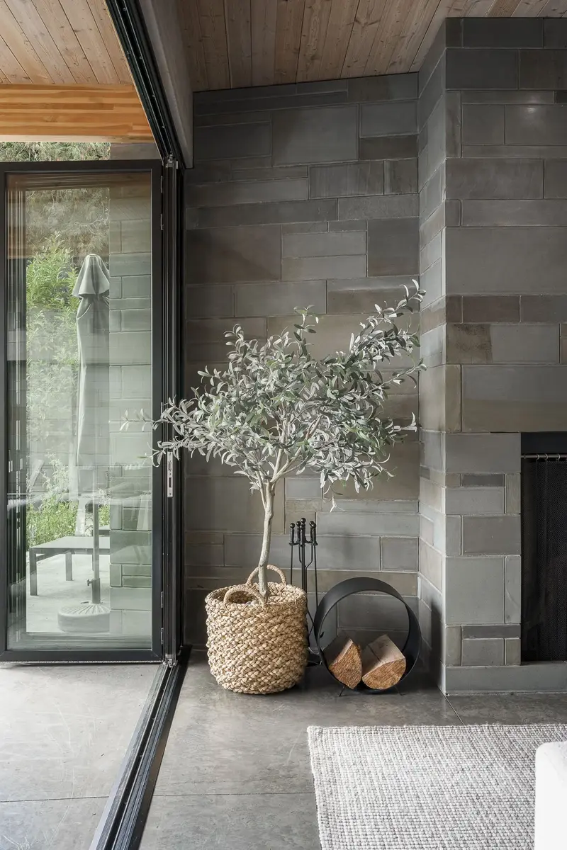 A stone fireplace beside a glass slider, a potted tree and stacked firewood alongside