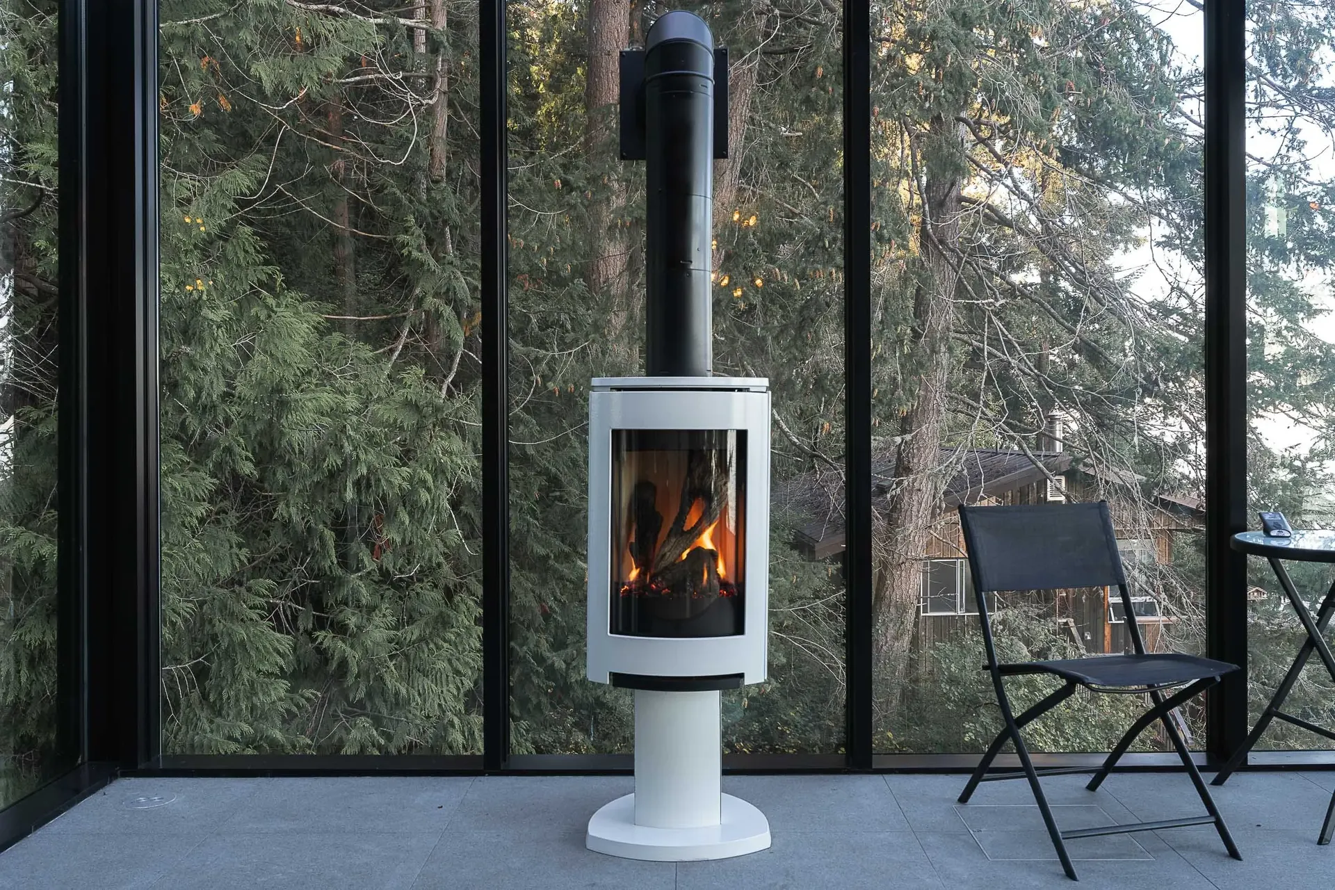 Freestanding white stove alight in front of full height glazing onto the trees