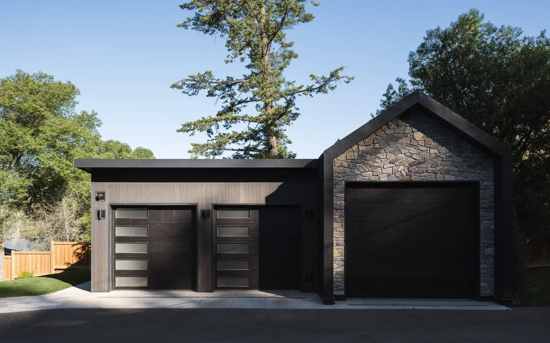 Three garage doors under a low dark roof beside a stone gable, seen from the driveway