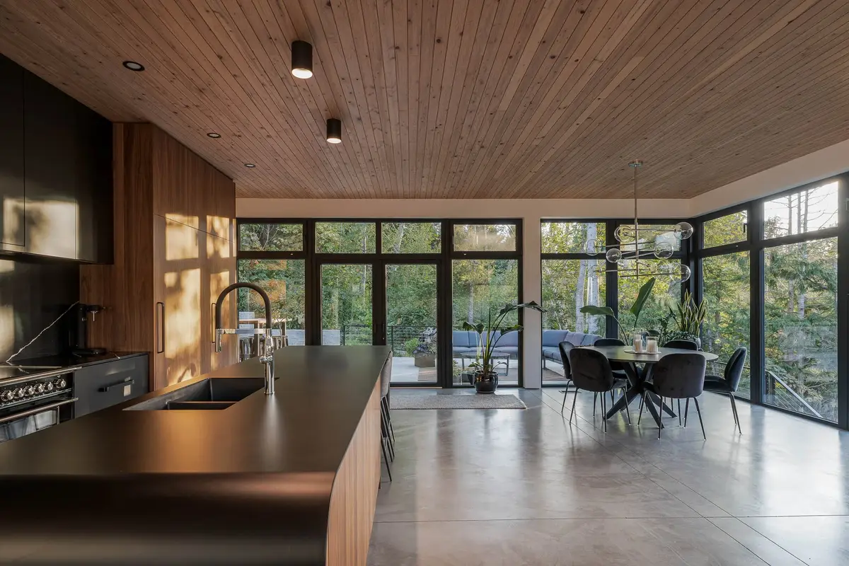Black range and a long dark island under a wood plank ceiling, glazing wrapping the dining area