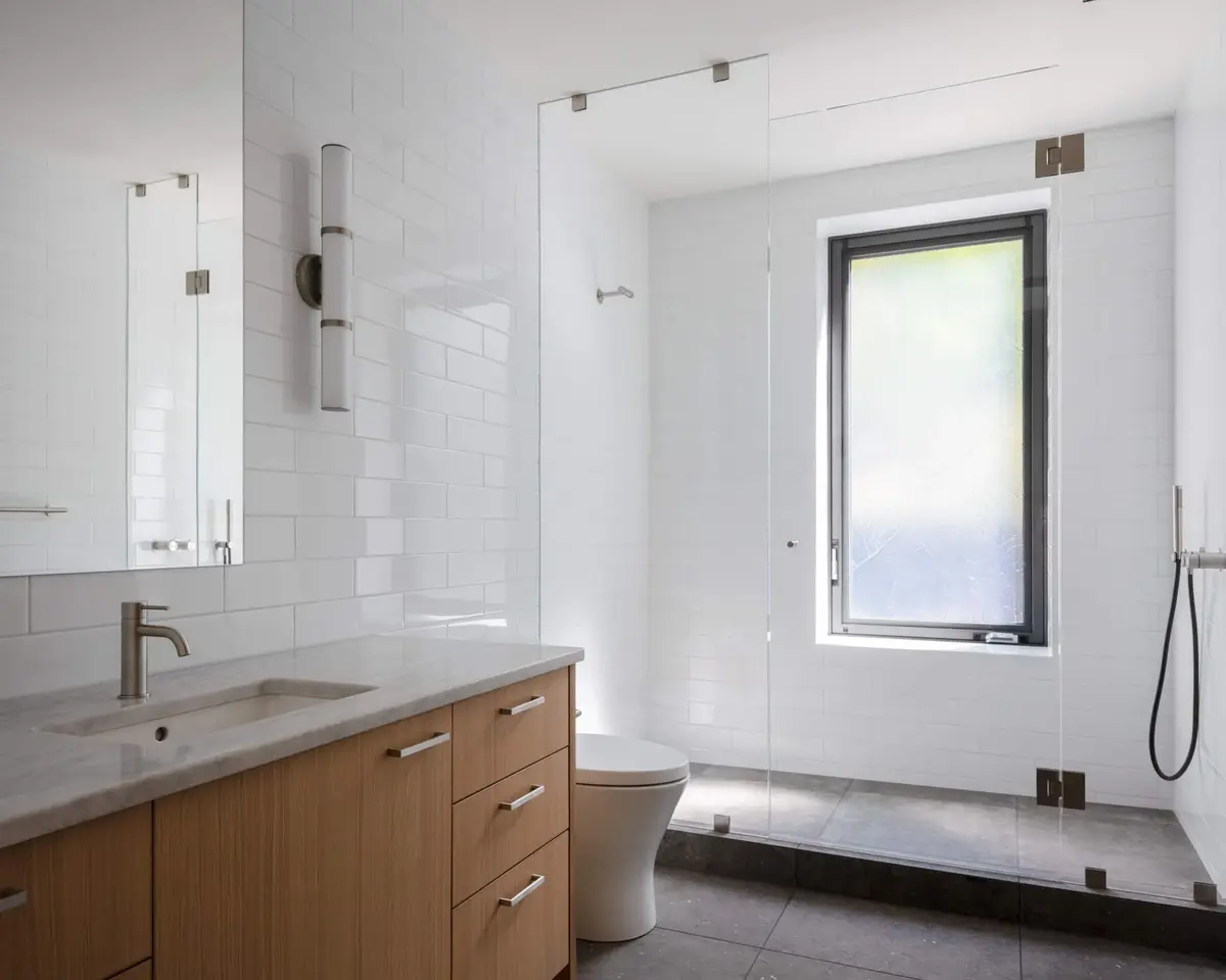 White tiled bathroom with a wood vanity, a glass shower and a window to the trees