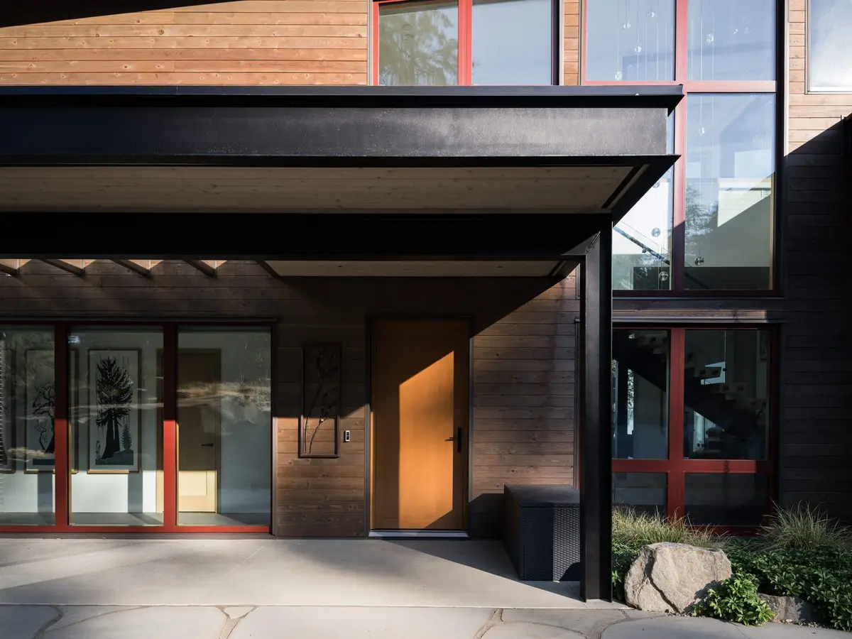 Timber front door under a dark steel canopy, red framed windows to either side