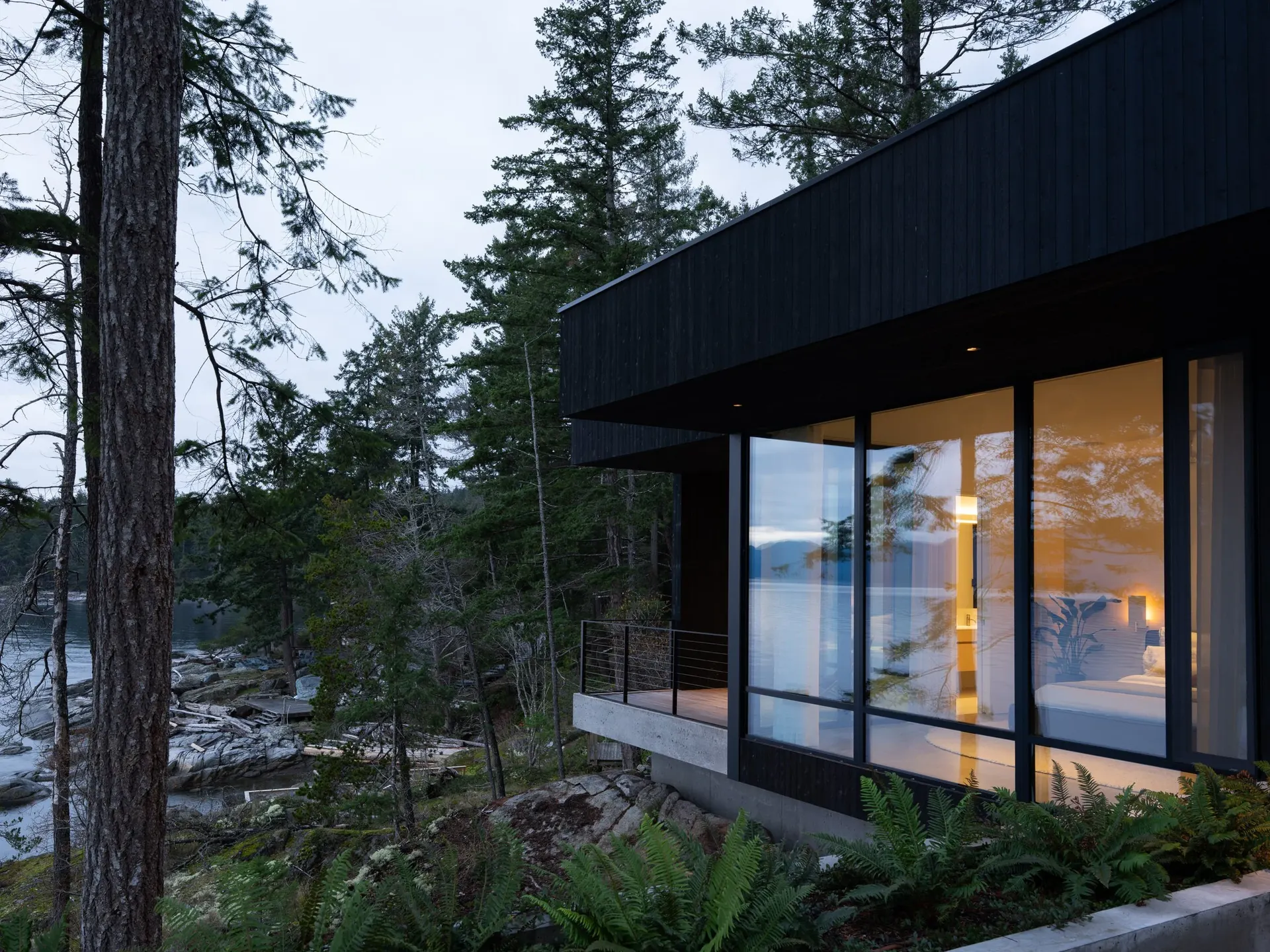 Dusk, a lit bedroom behind glass above a concrete base with ferns across the rock