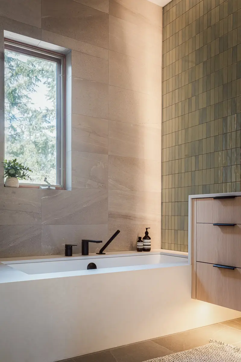 Tub below a window, green tile meeting the pale vanity beside it