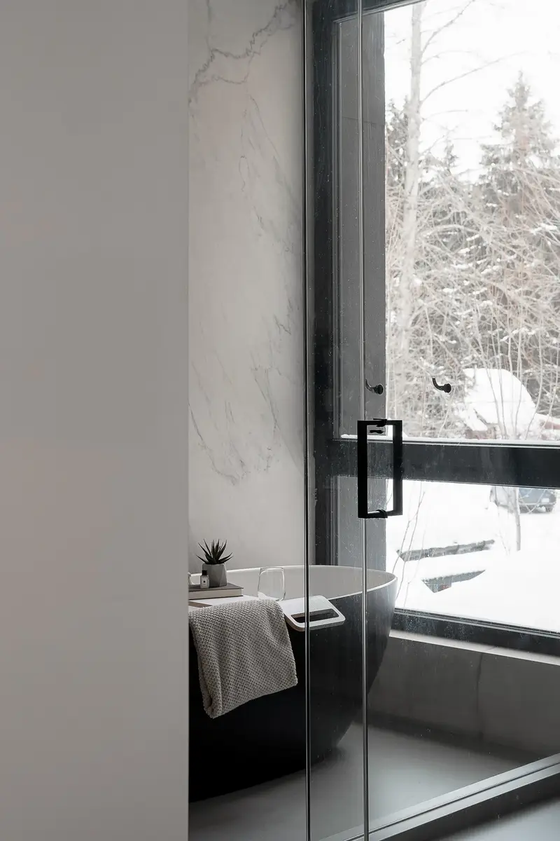Freestanding tub at a full height window, veined stone wall and a glass shower door