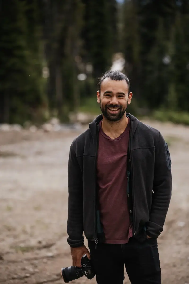 Matt Anthony standing on a gravel clearing holding a camera, conifers behind him