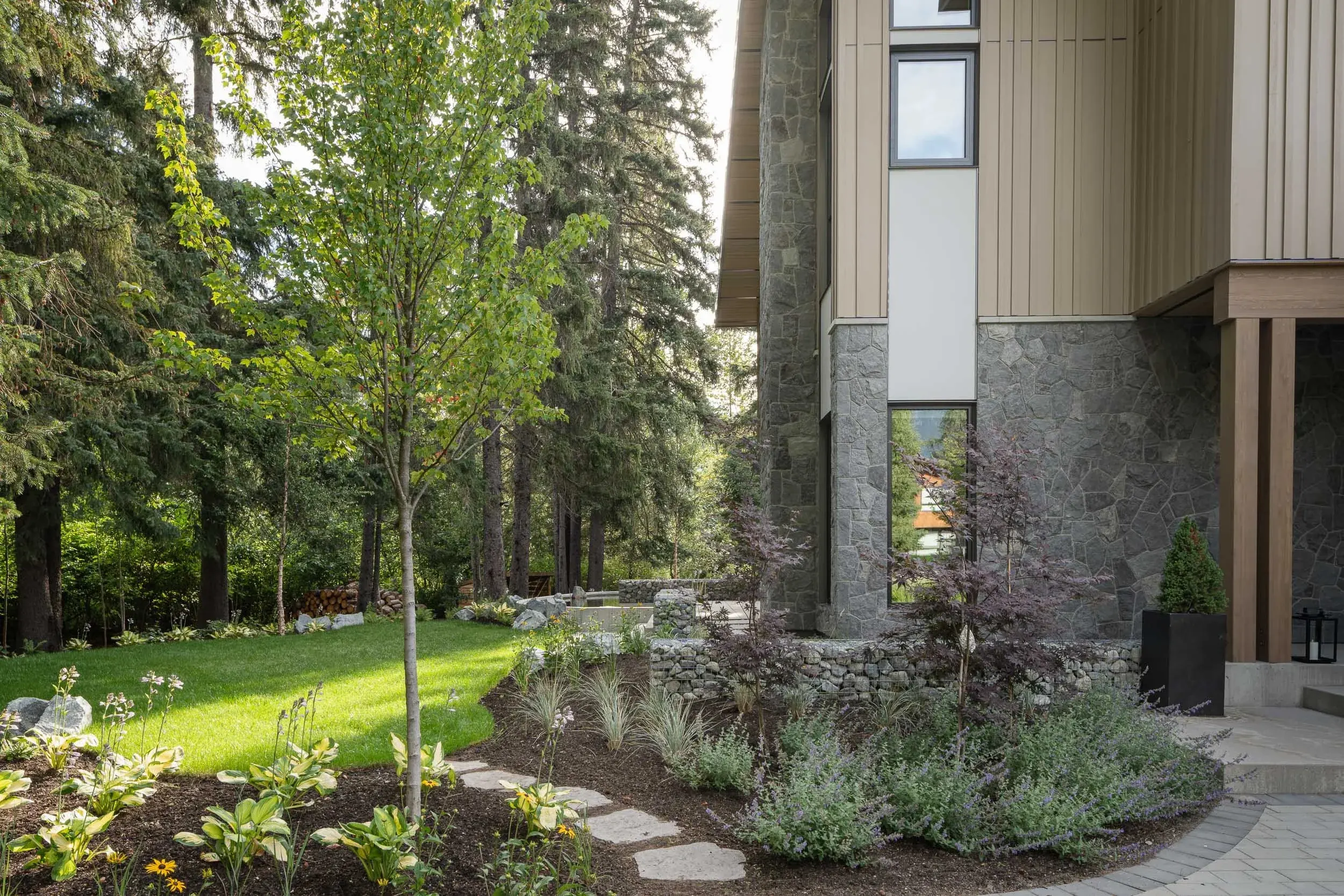Lawn and planted beds below a house with a stone base and pale vertical cladding
