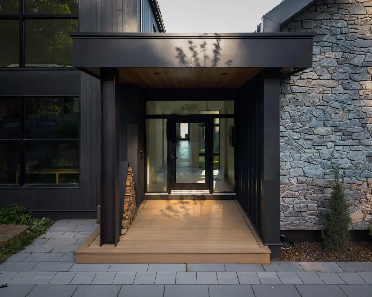 Covered entry with a wood soffit and stacked firewood, glass door open to a lit hall