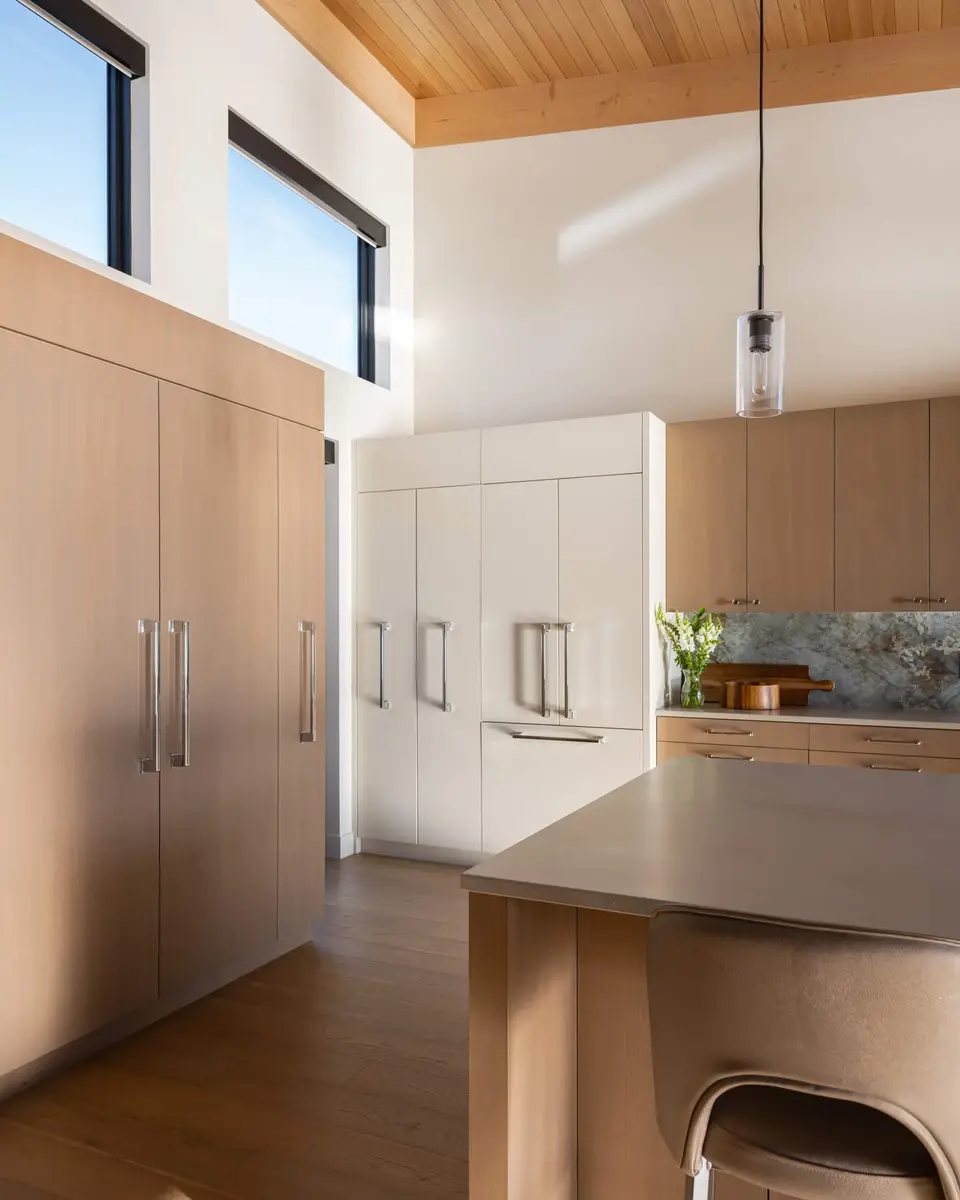 Tall pale wood and white cabinetry under high windows, an island edge and stool at right