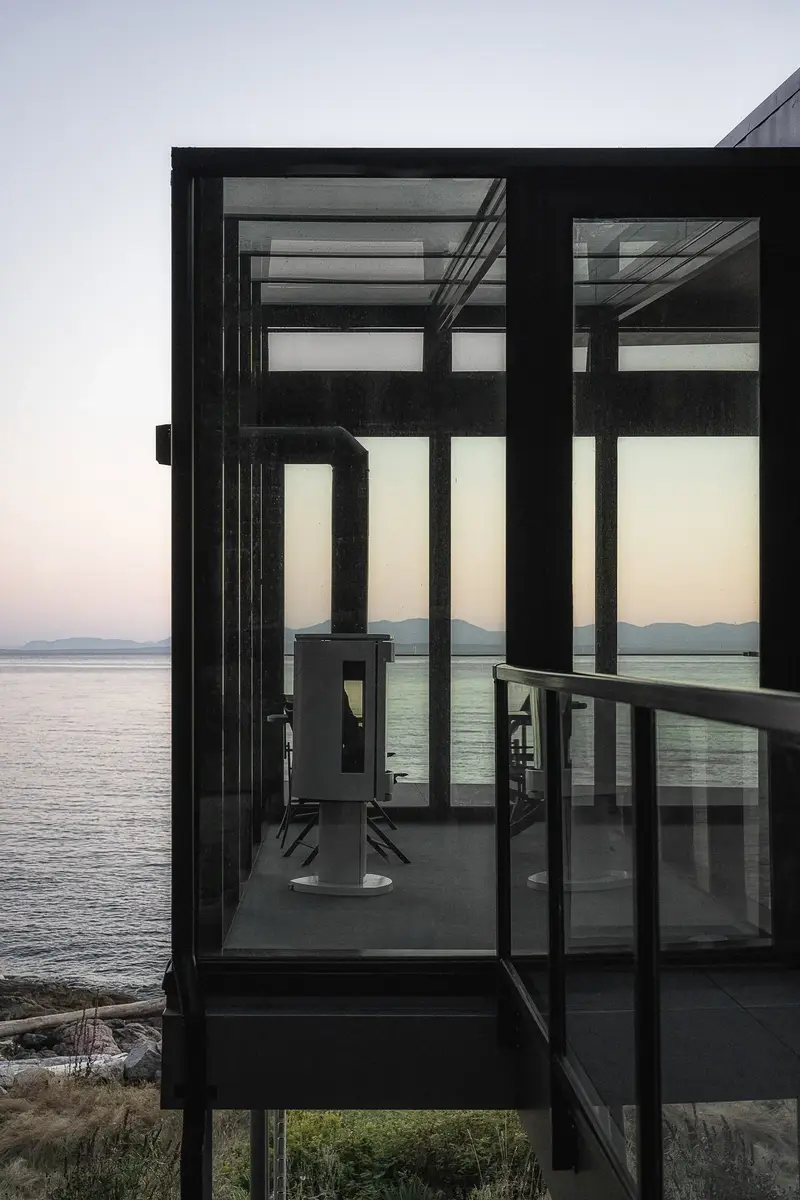 Glazed corner room at dusk above the shore, a stove inside and the sea beyond