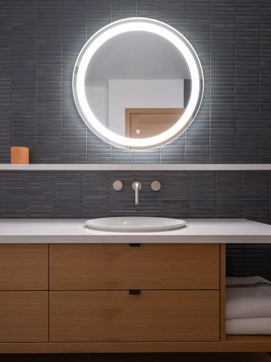 White basin under a round lit mirror, dark ribbed tile above pale timber drawers