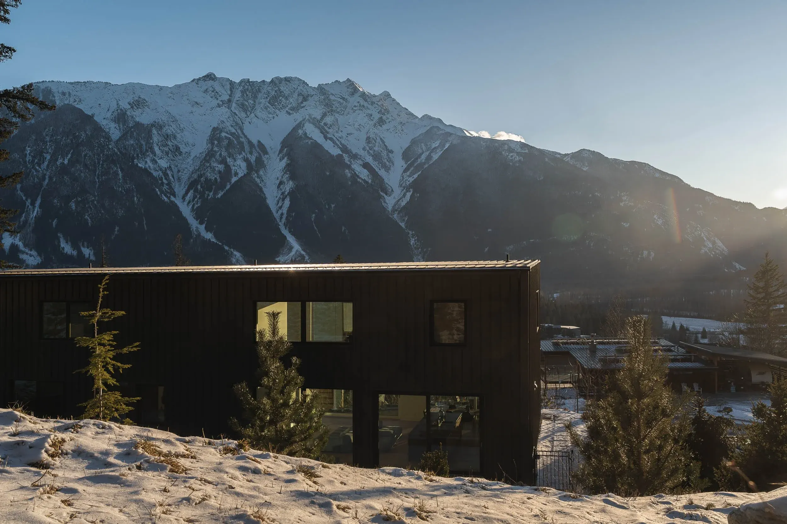 The house low on a snowy slope, a long dark elevation with snow covered peaks beyond