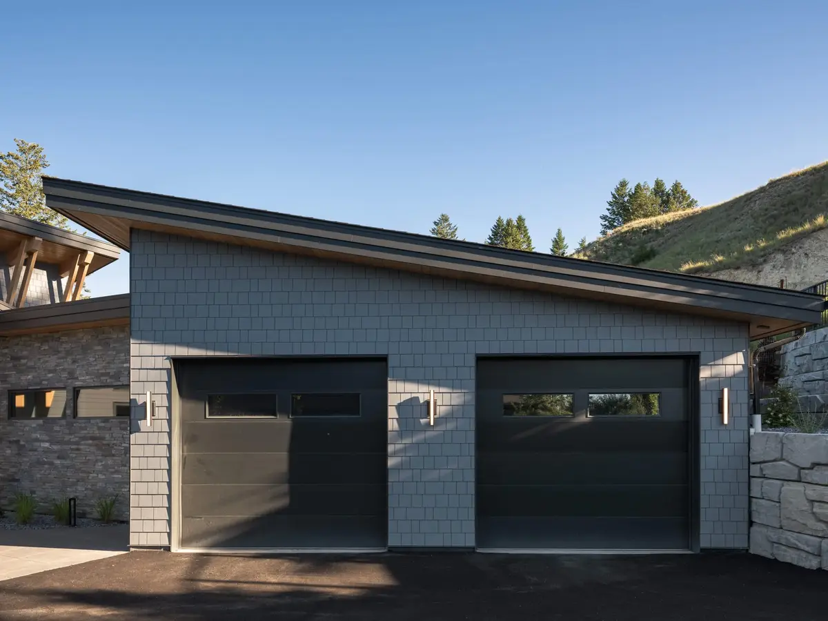Detached garage clad in shingles with two dark doors, a stone base and hillside behind