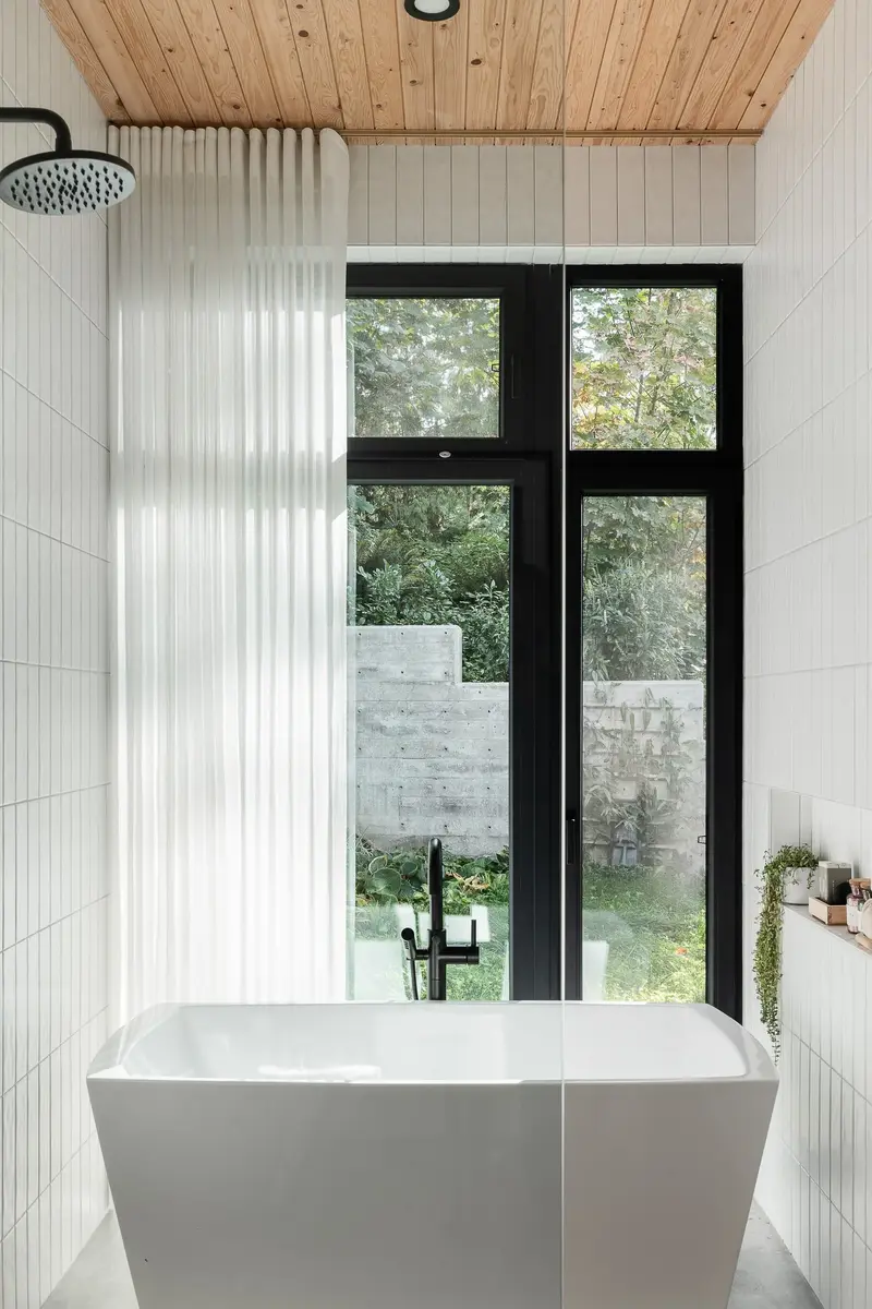Freestanding tub at a tall window, a sheer curtain beside it and a concrete wall outside