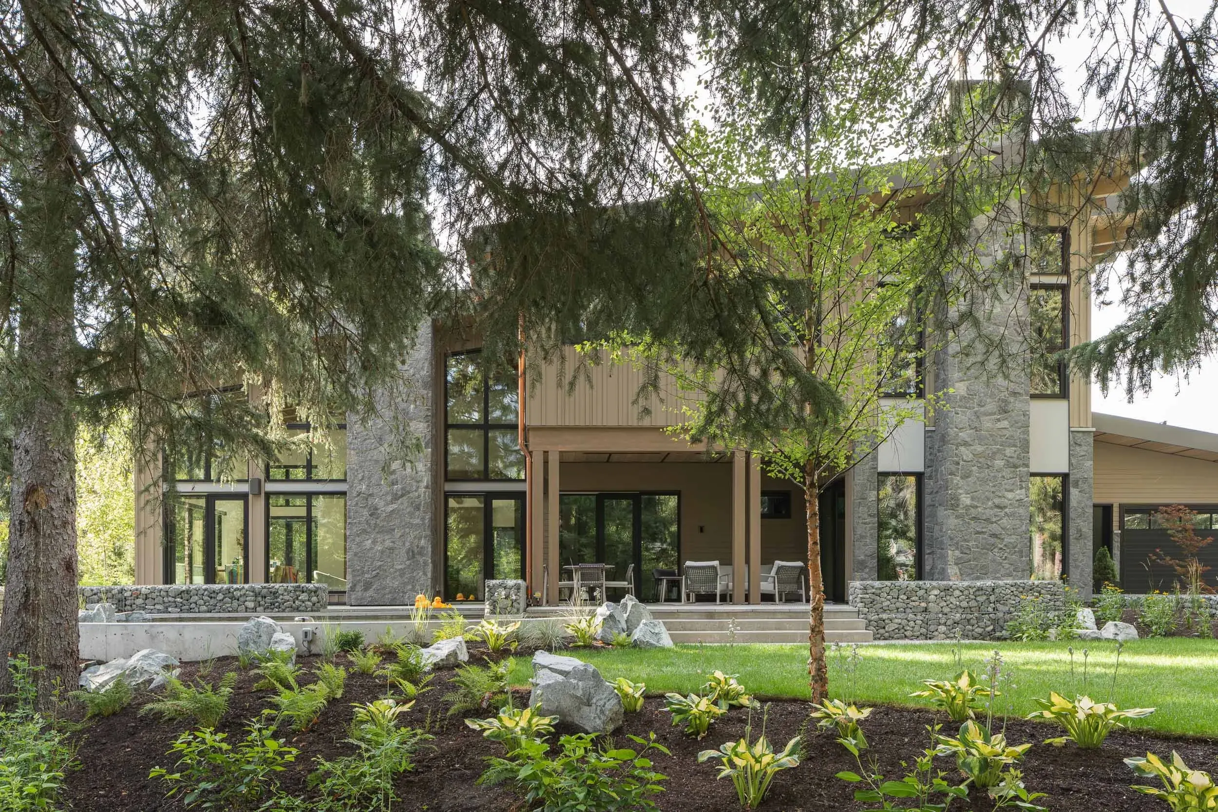 The house across a lawn seen under a conifer bough, stone base and glazed living rooms