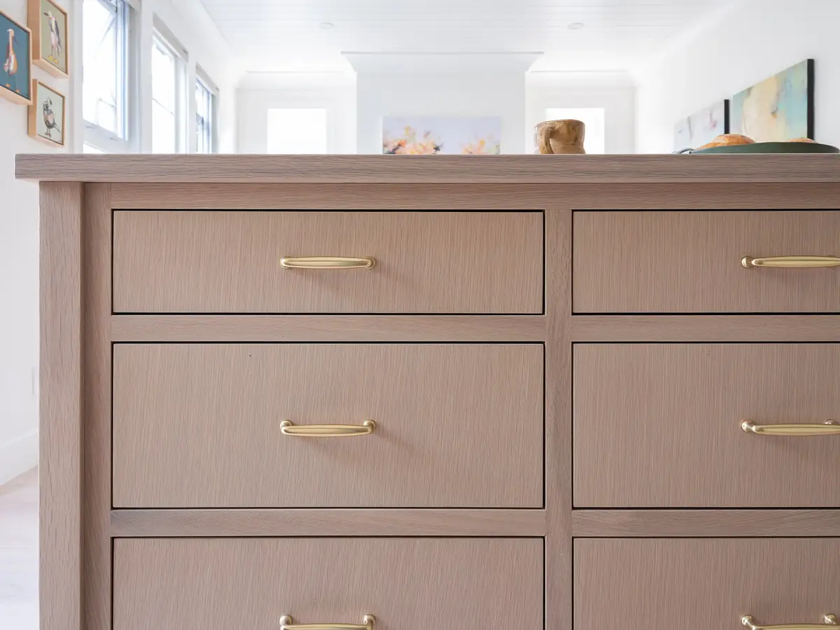 Island drawer fronts in pale wood with slim gold pulls, the room soft behind