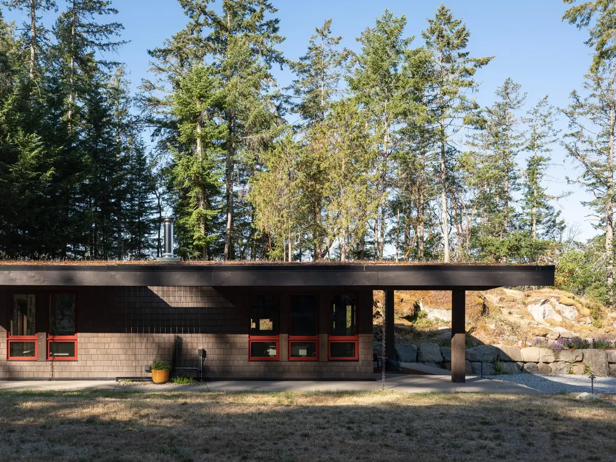 Low outbuilding under a planted roof, red framed windows across a dark shingled wall