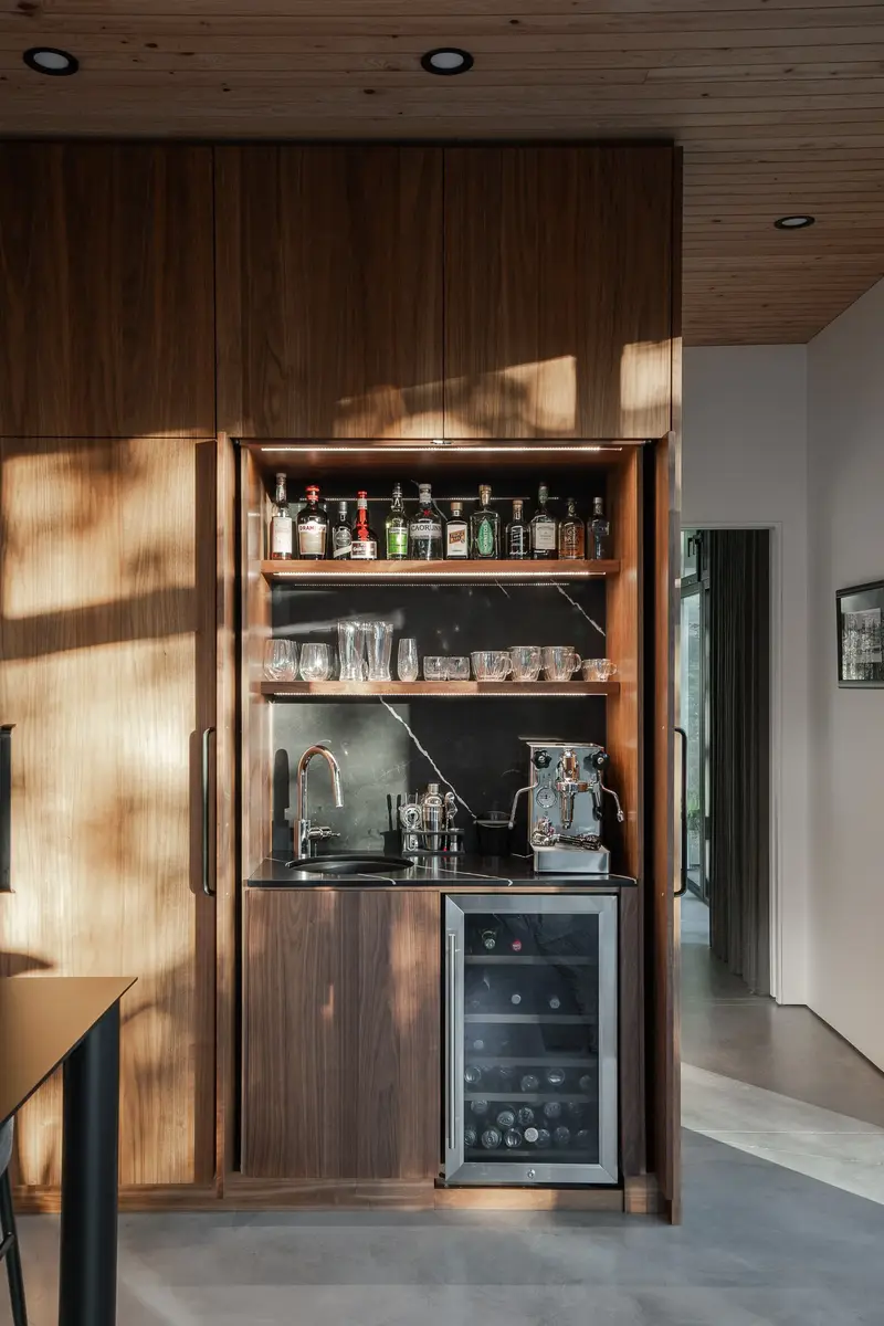 Bar behind sliding wood doors, lit shelves of bottles and glassware above a sink and wine fridge