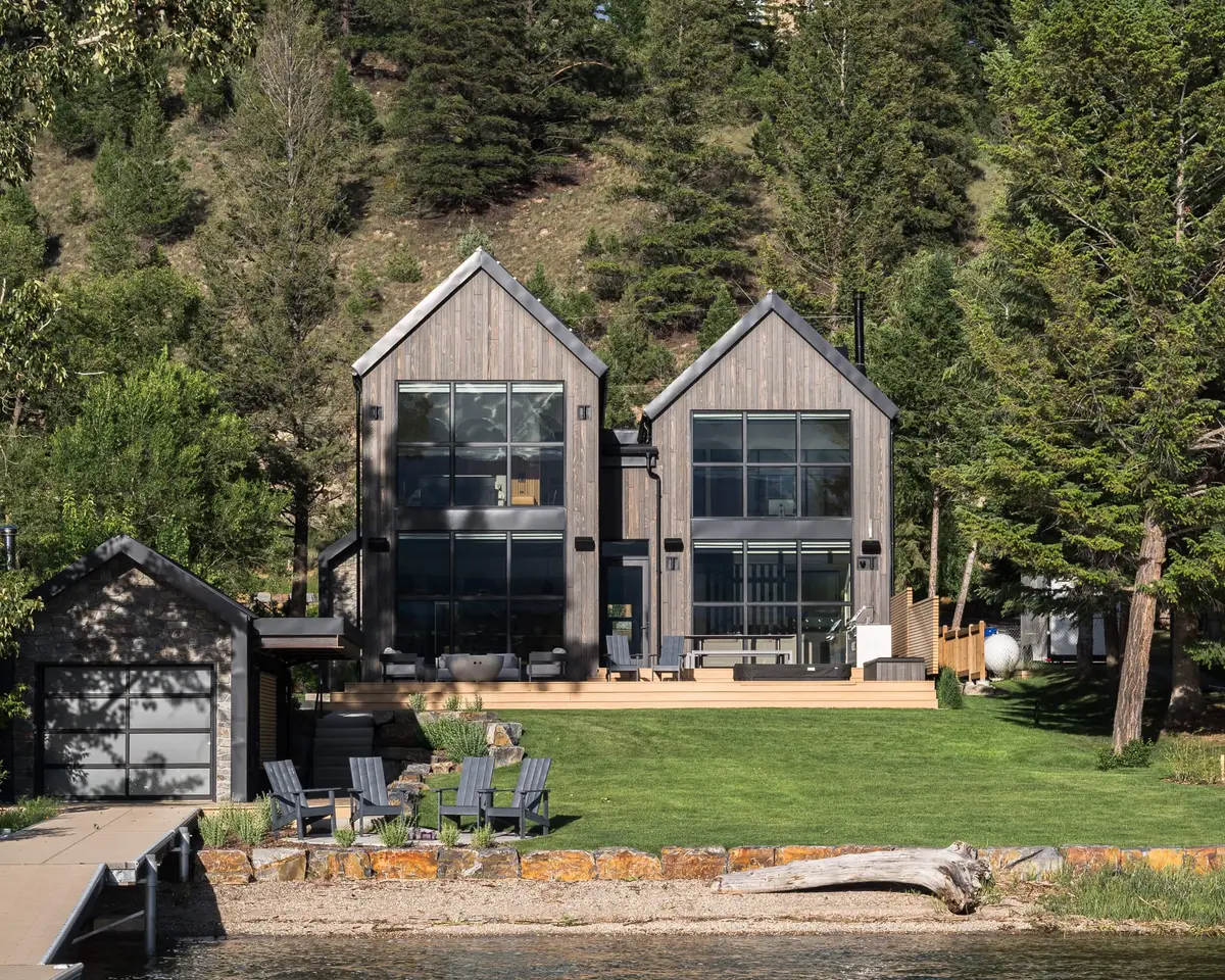 Twin gabled volumes in weathered wood above a lawn, seen from the dock across the shoreline