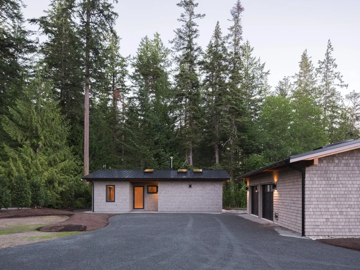 Gravel drive between two outbuildings, lit sconces under the eaves and tall trees behind