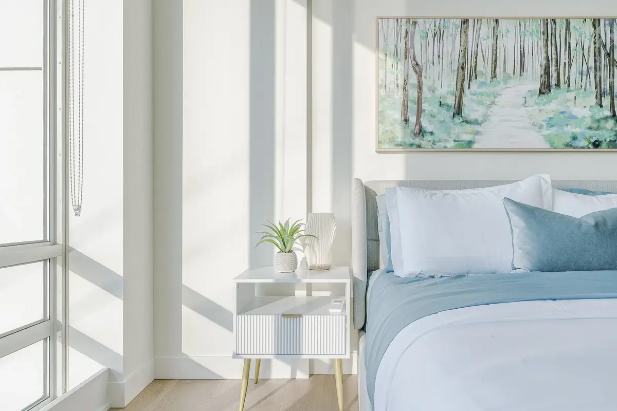 Sunlight falling across a bedside table, blue linens and a small plant beside the lamp