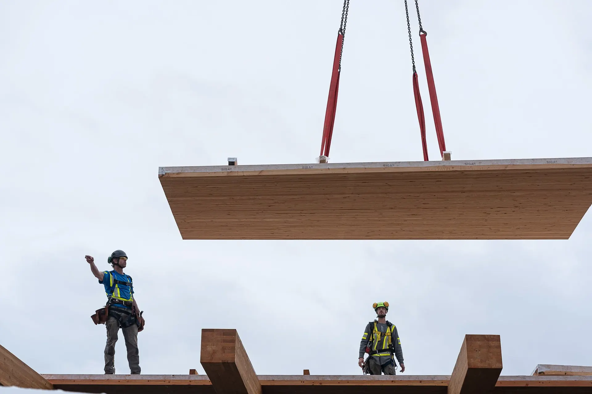 Two workers on a mass timber frame, a panel hanging from the crane