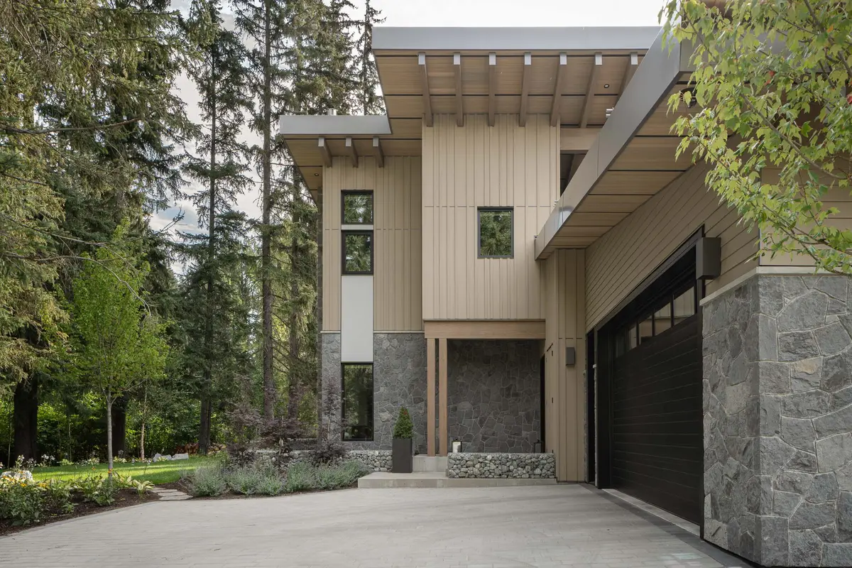 Entry and garage in spring, a wood soffit on exposed beams, a dark garage door, and a stone pier
