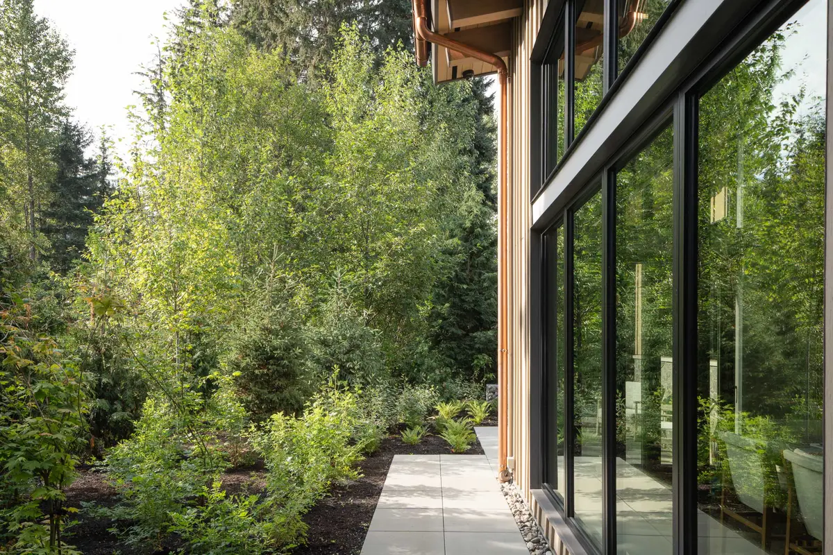 A copper downspout beside vertical wood and black framed glass, a stone path and young trees