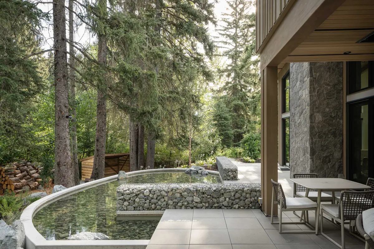 A curved pool edged in gabion beside the terrace, a table and chairs under the wood soffit, firs behind