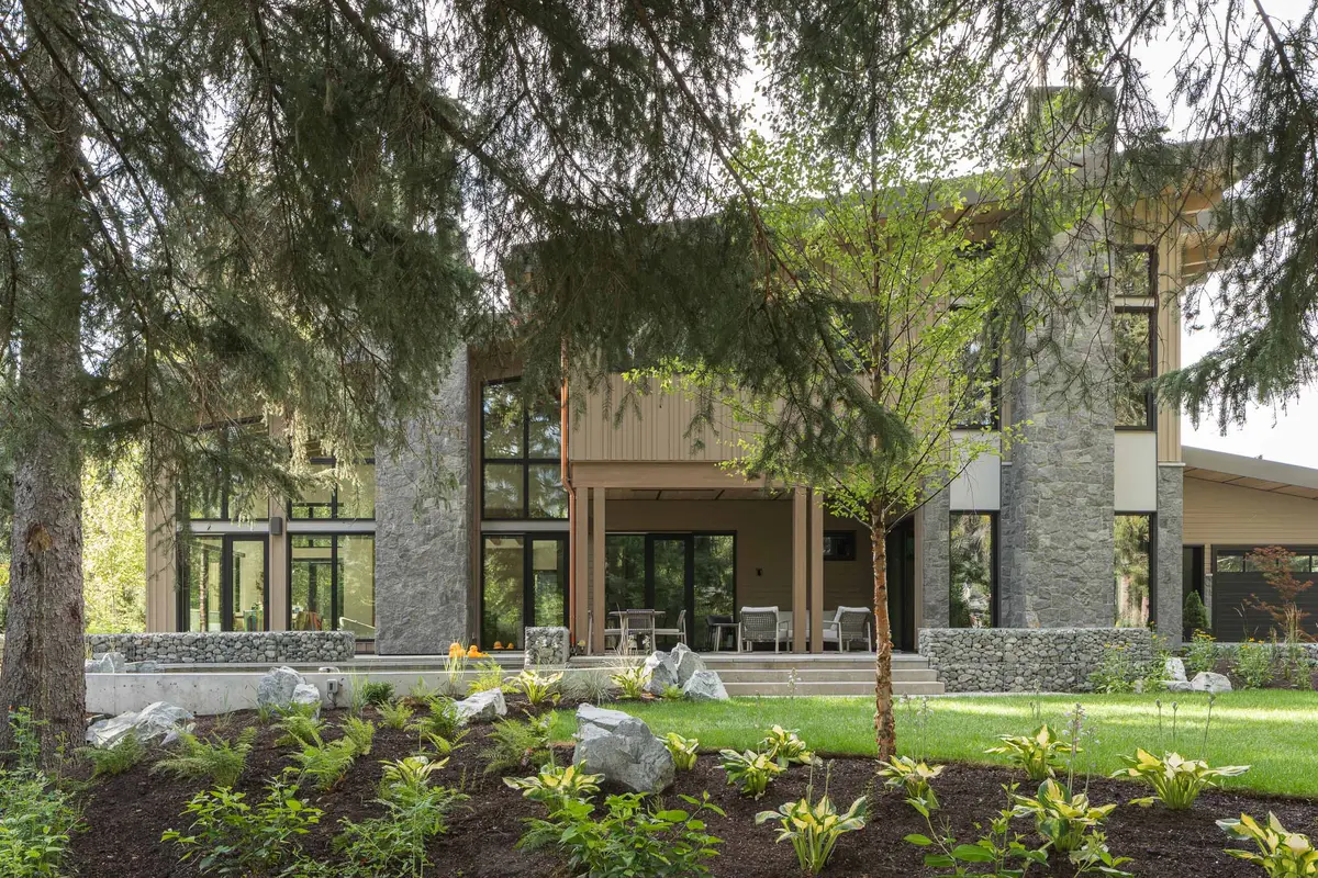 Rear elevation in spring, stone piers and tall glazing seen through fir branches, with a covered seating area and a lawn